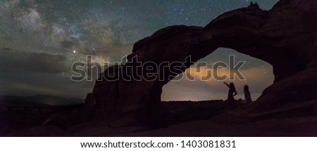 Two women enjoying each other's company while taking in the Milky Way under Broken Arch in Arches National Park. A low flying moon illuminates the clouds in the bottom of the photo.