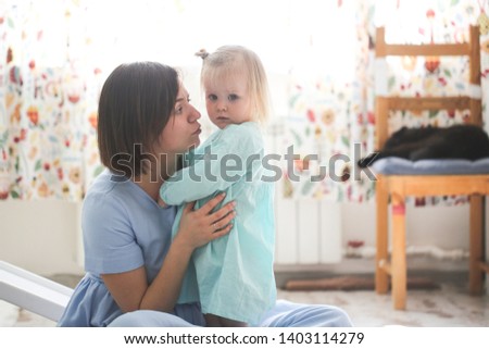 cute girl babe hugs mom, daughter toddler blonde with fluffy hair in her mother's arms in a bright real room, the concept of a safe childhood, affection and emotions