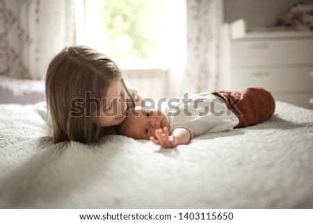 The older sister kisses the baby for 1 month, the girl embraces the newborn baby on the bed in the bedroom. Against the background of the window in the real bedroom