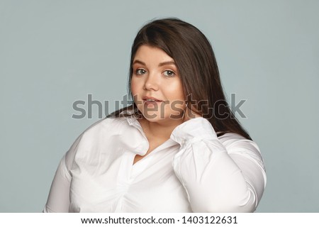 Picture of stylish young overweight female employee wearing white shirt and large round earrings touching her neck. Neat beautiful chubby woman posing against gray scopyspace tudio wall background