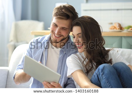Young couple watching media content online in a tablet sitting on a sofa in the living room. Royalty-Free Stock Photo #1403160188