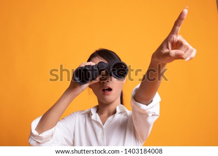 Young Asian woman  point and look with binoculars on yellow background