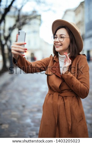 Gorgeous young woman waving hand on city background during video call. Outdoor photo of enthusiastic girl with wavy hairstyle making selfie while sitting on the street in european country.