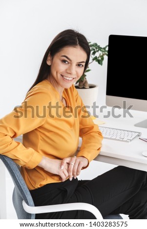 Photo of joyful brunette businesswoman 20s sitting at table and working on big computer in bright office