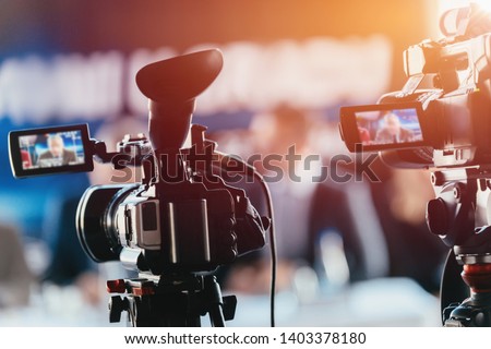 Two cameras recording presentation at press conference, blurred speakers wearing suits background, live streaming concept Royalty-Free Stock Photo #1403378180