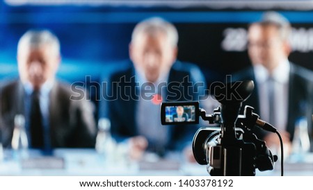Professional digital camera recording presentation of a blurred speakers wearing suits, live streaming concept