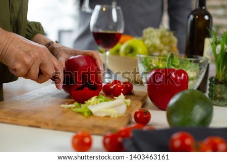 Cutting red pepper. Close-up picture of aged wrinkled caring loving female wearing elegant emerald dress cutting red peeper for the dinner