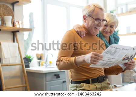 Embracing a spouse. Smiling attractive glowing handsome silver-haired bearded man in a mustard sweater engaging in reading a paper while his tender aging white-haired spouse embracing him.