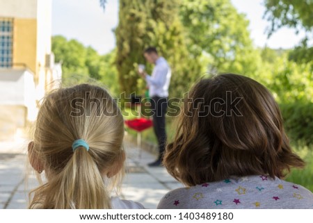Two small girls are watching a performance of a magician through an outdoor birthday party .