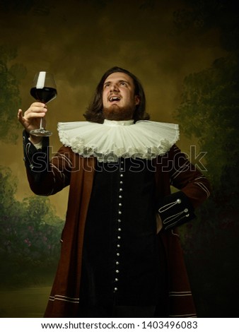 Young man as a medieval knight on dark studio background. Portrait of male model in retro costume. Holding a glass of red wine. Human emotions, comparison of eras, facial expressions concept.