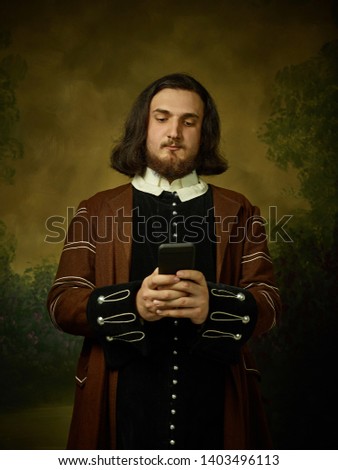 Young man as a medieval knight on dark studio background. Portrait in low key of male model in retro costume. Making selfie. Human emotions, comparison of eras and facial expressions concept.