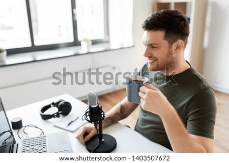 technology, mass media and podcast concept - happy male audio blogger with laptop computer and microphone drinking coffee and broadcasting at home office