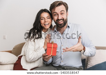 Image of charming couple man and woman holding birthday present box while sitting on sofa in bright room at home