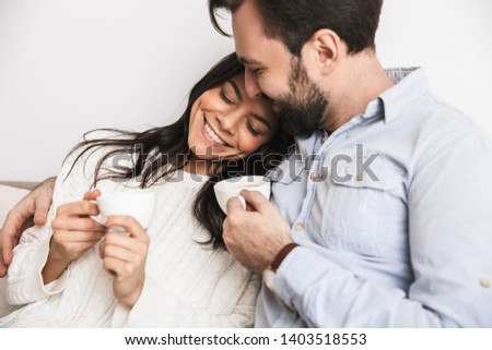 Image of caucasian brunette couple man and woman drinking coffee together while sitting on couch in bright room at home