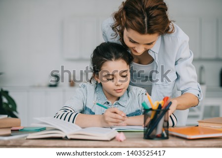 adorable child writing in copy book while doing schoolwork near mother Royalty-Free Stock Photo #1403525417