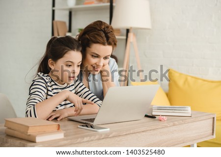 surprised mother and daughter using laptop while doing homework together Royalty-Free Stock Photo #1403525480