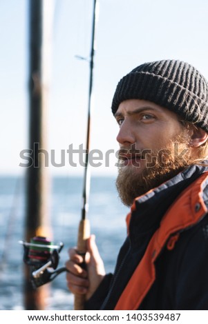Handsome brutal bearded fisherman wearing coat standing with a fishing rod at the seashore