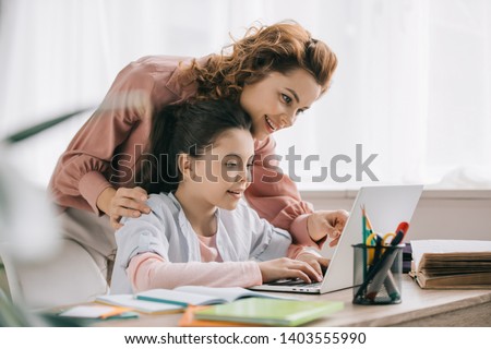 happy mother helping smiling daughter doing schoolwork at home Royalty-Free Stock Photo #1403555990
