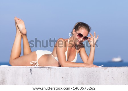 Girl resting on concrete breakwater. Attractive young woman in sunglasses lies on front on concrete wall and looking at camera through sunglasses