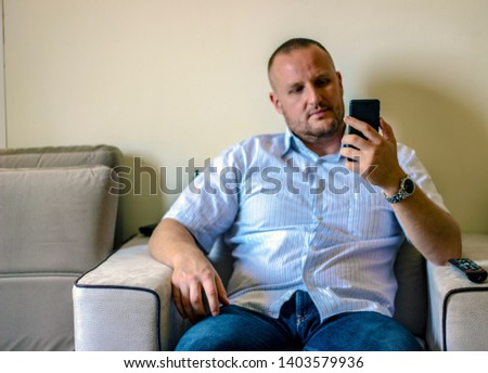 Man sitting relaxed on sofa and checking his smartphone messages.  Happy young man using modern smartphone device while sitting on sofa at home, modern design interior. Focus on hand with phone.