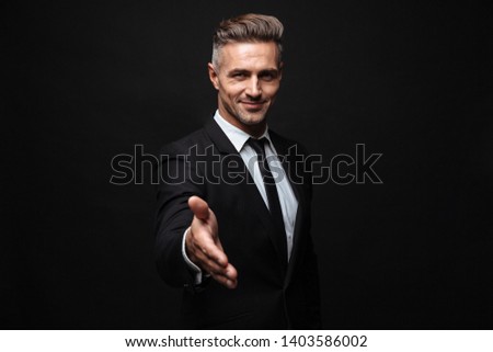 Confident attractive businessman wearing suit standing isolated over black background, outstretched hand fir greeting