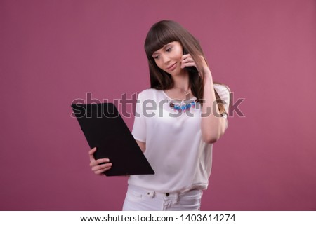 Image of young business woman standing over pink background looking aside chatting by phone holding folder.