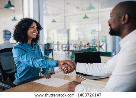Smiling African American manager sitting at his desk in an office shaking hands with a potential new employee after an interview Royalty-Free Stock Photo #1403726147
