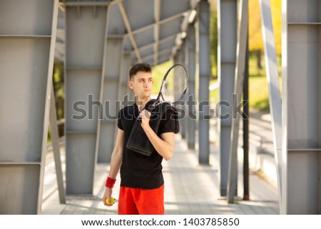 The man poses outdoors with a tennis racket and a ball. A towel is hanging on his shoulder. Sport concept