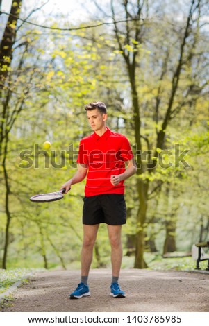 The man in a red t-shirt pose with a tennis racket and a ball on the background of green park. Sport concept