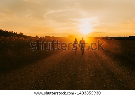 Beautiful couple in love is walking the road between the fields of sunflowers, summer sunset, bright orange light. In the background is a forest and rustic wooden houses. Concept of travel by car, eco