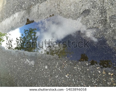 A reflection of blue sky, white clouds and branches with green leaves, in a concrete bar