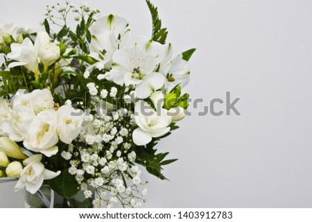 Home decoration still life of delicate white spring flowers in transparent glass vase standing on a shelf against white wall.