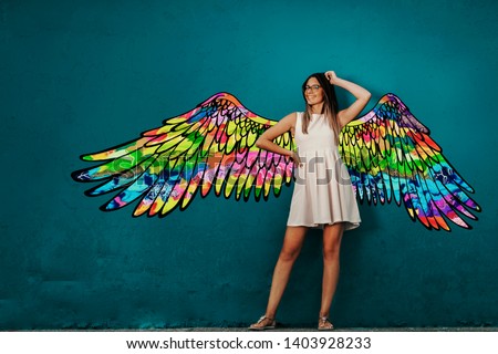 Smiling Caucasian girl in white summer dress posing in front of turquoise wall with colorful wings. Royalty-Free Stock Photo #1403928233