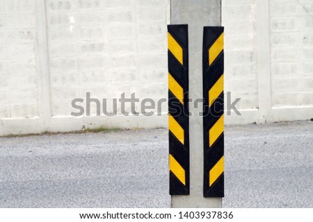 warning striped road yellow-black on both side of electrical concrete pole a black car passing on road thru back in the public car park