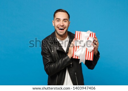 Handsome stylish young bearded man in black leather jacket white t-shirt hold gift box isolated on blue wall background studio portrait. People sincere emotions lifestyle concept. Mock up copy space