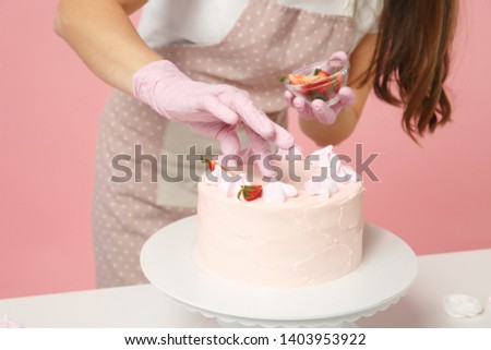 Close up cropped chef cook confectioner baker in apron white t-shirt cooking at table isolated on pink pastel background in studio. Cake decorating process strawberry meringue. Mock up food concept