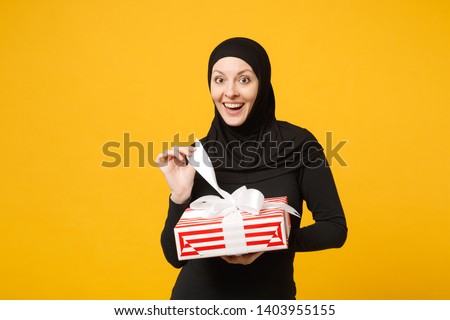 Smiling young arabian muslim woman in hijab black clothes hold in hand present box with gift isolated on yellow wall background studio portrait. People religious lifestyle concept. Mock up copy space