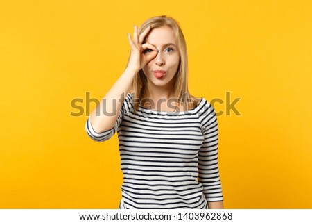 Funny young woman in striped clothes holding hand near eyes imitating glasses or binoculars showing tongue isolated on yellow orange background. People emotions, lifestyle concept. Mock up copy space