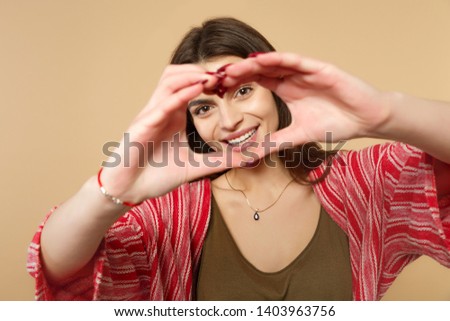 Portrait of smiling young woman in casual clothes showing shape heart with hands isolated on pastel beige wall background in studio. People sincere emotions, lifestyle concept. Mock up copy space.