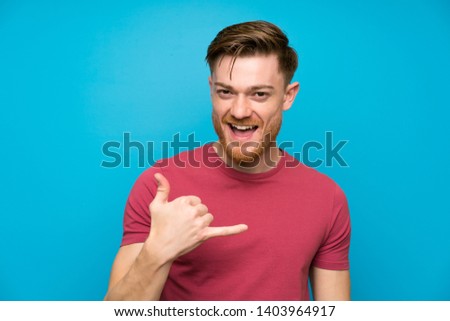 Redhead man on isolated blue wall making phone gesture