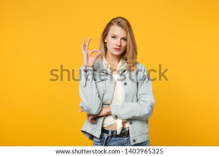 Portrait of attractive confident young woman in denim casual clothes looking camera, showing OK gesture isolated on yellow orange background in studio. People lifestyle concept. Mock up copy space