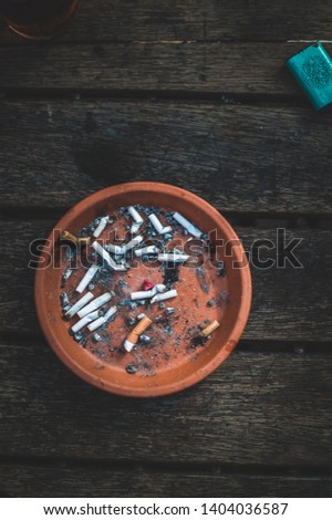 Close up picture of ashtray, full uf cigarettes and ashes. Dark desaturated colours, sad mood. Bad habits. Dark brown real wood table, Bright orange ashtray, dirty habit. Bad smell.