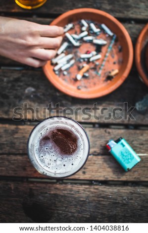 Close up picture of an ashtray full of smoked cigarettes. No brand Lighter and ice cold pint of nice beer. Hand in a shot, using an ashtray, smoking person, siting in a bar. Wooden table, natural.