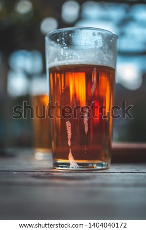 Close up picture of ice cold pint of tasty beer, amber colour, big nice glass, wooden table, blue sky, trees and sun. Natural light, natural colours. Alcoholic drink just for adults. Good time.
