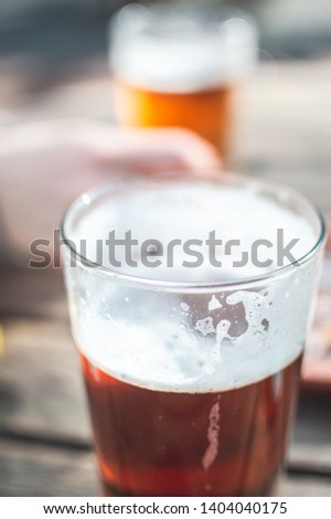 Close up picture of ice cold pint of tasty beer, amber colour, big nice glass, wooden table, blue sky, trees and sun. Natural light, natural colours. Alcoholic drink just for adults. Good time.