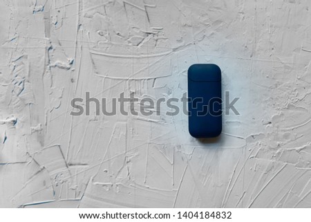 Closed blue IQOS lies in the right of the image on white decorative abstract plaster texture with textured smears. The new technology cigarette, hybrid cigarette, heatsticks, tobacco, new device.