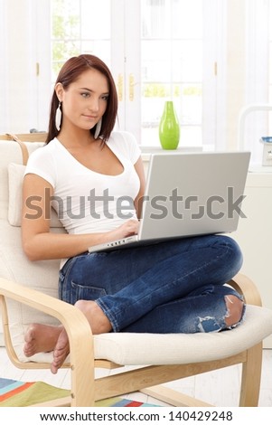 Young woman sitting in armchair typing on laptop computer at home.