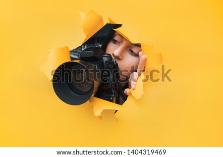 A photographer paparazzi girl with a black DSLR camera looks at what is happening from the ambush with curiosity. Yellow paper, torn hole. Tabloid press. In search of the plot for photo stock.