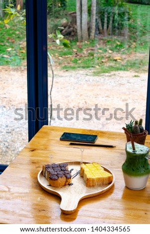 Iced cold Matcha green tea and milk layer drink in a jar with straw and crispy toasted snack bread with honey, chocolate, and sugar on the wooden table next to the smartphone and pencil.