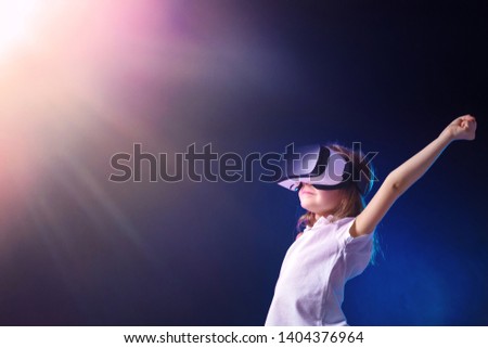 Excited female child using digital glasses of virtual reality rises hands up from happiness on shining blue background